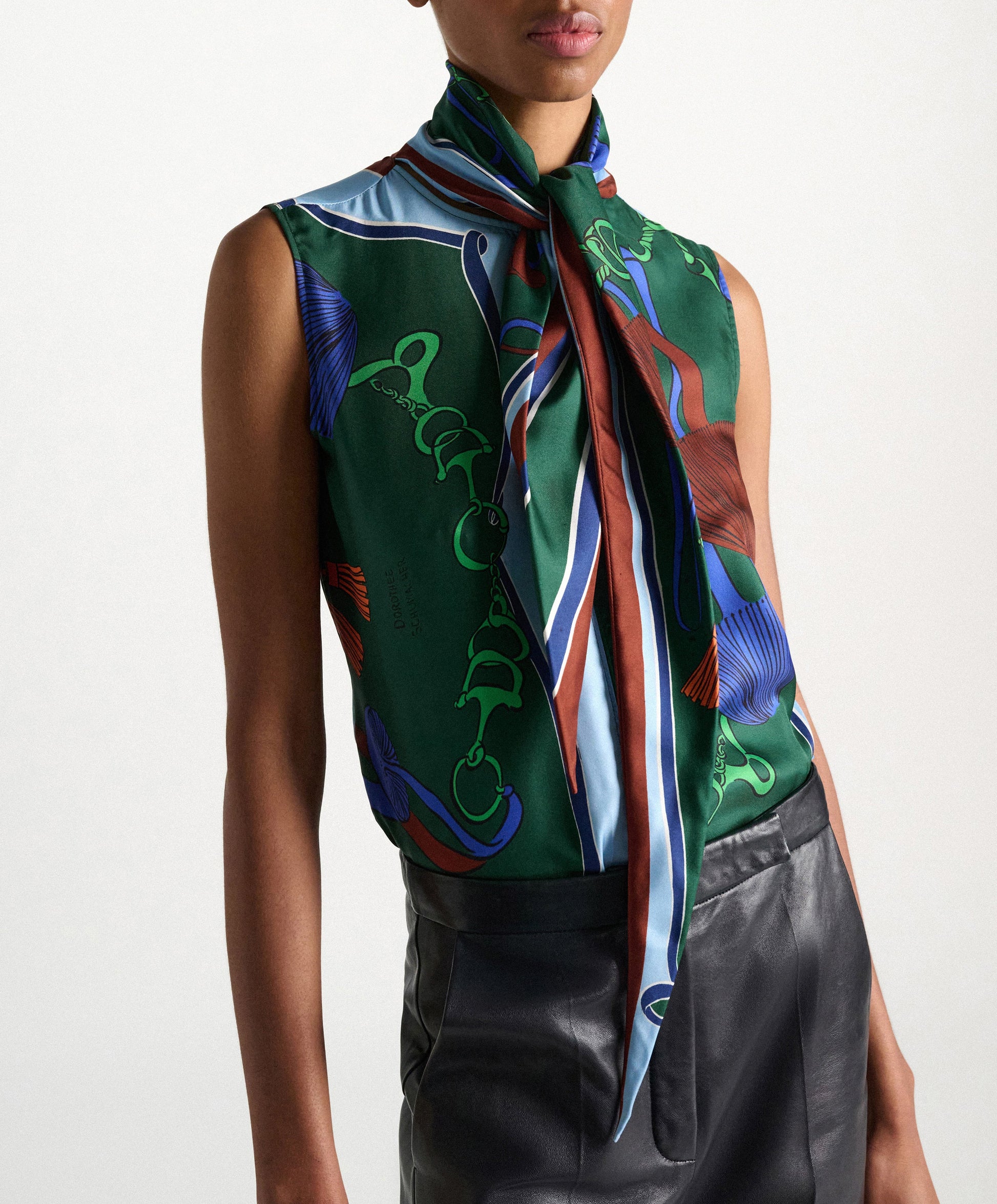 A person is wearing the Dorothee Schumacher Cosmic Funghi Top, a sleeveless blouse featuring a tie collar and a green printed pattern, paired with high-waisted black leather pants and shown from shoulders to hips.