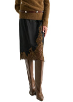 A person wearing a brown sweater, a brown belt with silver buttons, Dorothee Schumacher's Shiny Statement Skirt, and brown knee-high boots.
