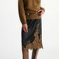 A person stands against a plain white background wearing the Dorothee Schumacher Shiny Statement Skirt—a black midi-skirt with delicate brown lace trim—styled with a fuzzy brown sweater, a brown waist belt, and tall brown boots.