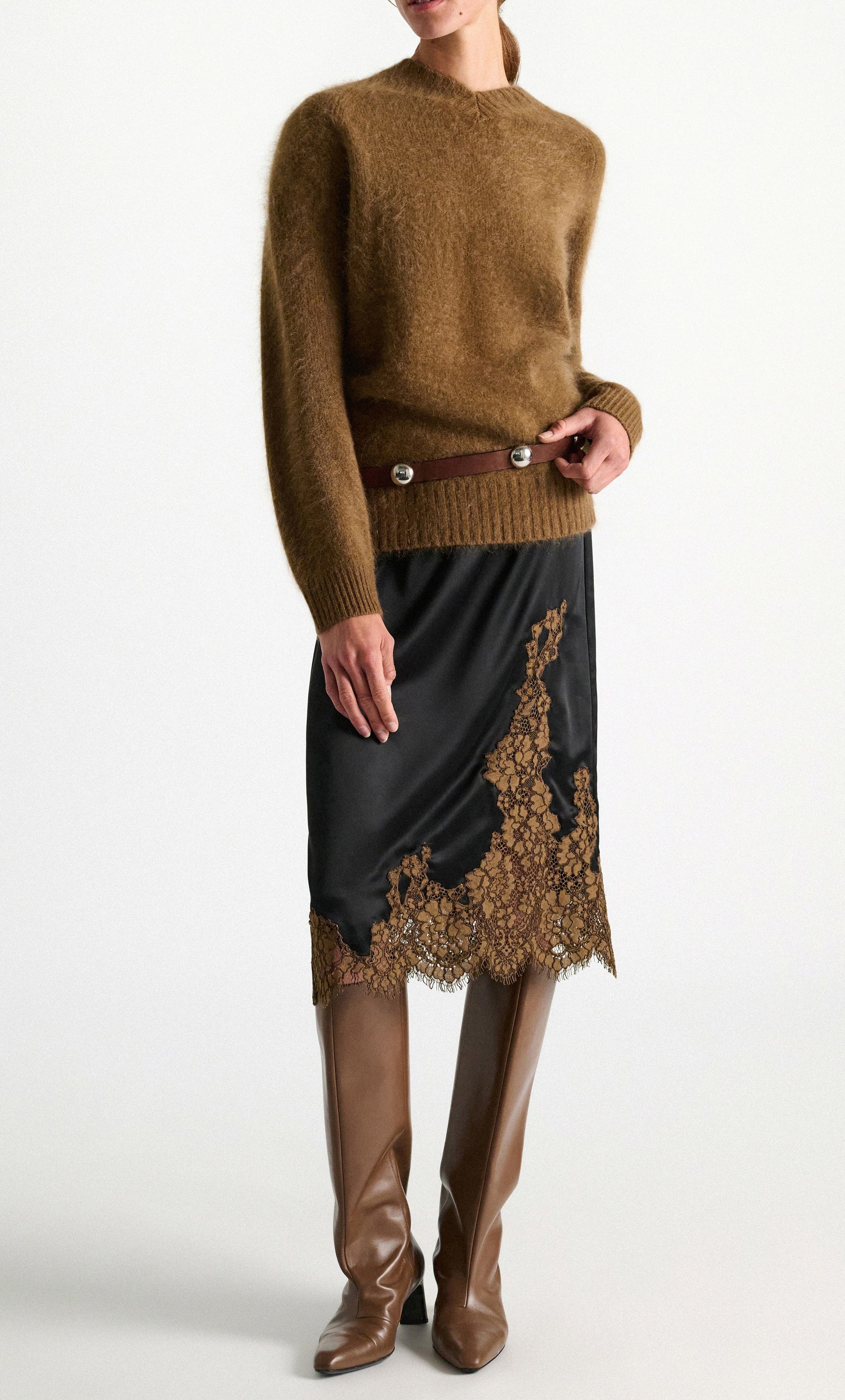A person stands against a plain white background wearing the Dorothee Schumacher Shiny Statement Skirt—a black midi-skirt with delicate brown lace trim—styled with a fuzzy brown sweater, a brown waist belt, and tall brown boots.