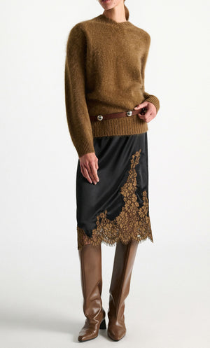 A person stands against a plain white background wearing the Dorothee Schumacher Shiny Statement Skirt—a black midi-skirt with delicate brown lace trim—styled with a fuzzy brown sweater, a brown waist belt, and tall brown boots.
