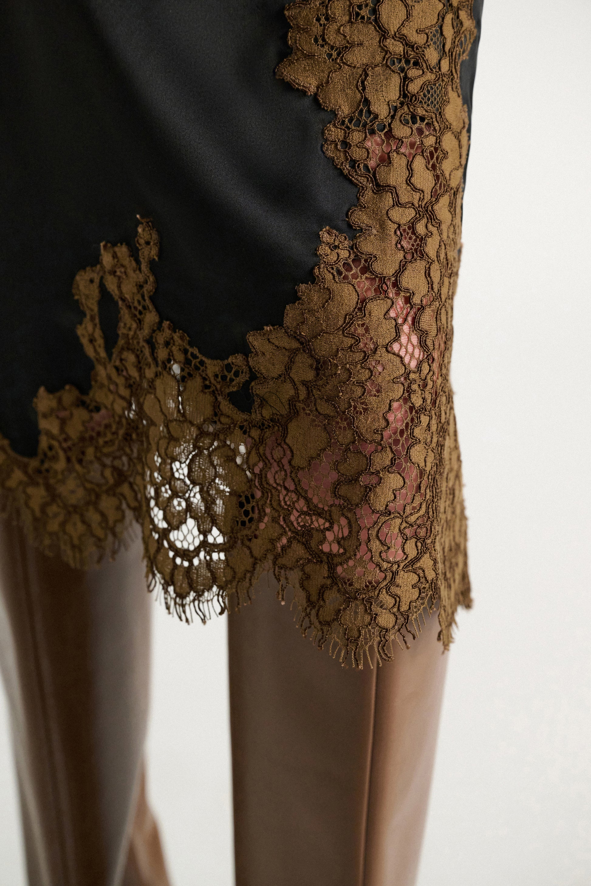 Close-up of the Dorothee Schumacher Shiny Statement Skirt, a black technical satin midi-skirt with brown lace trim at the hem, styled over brown leather pants.