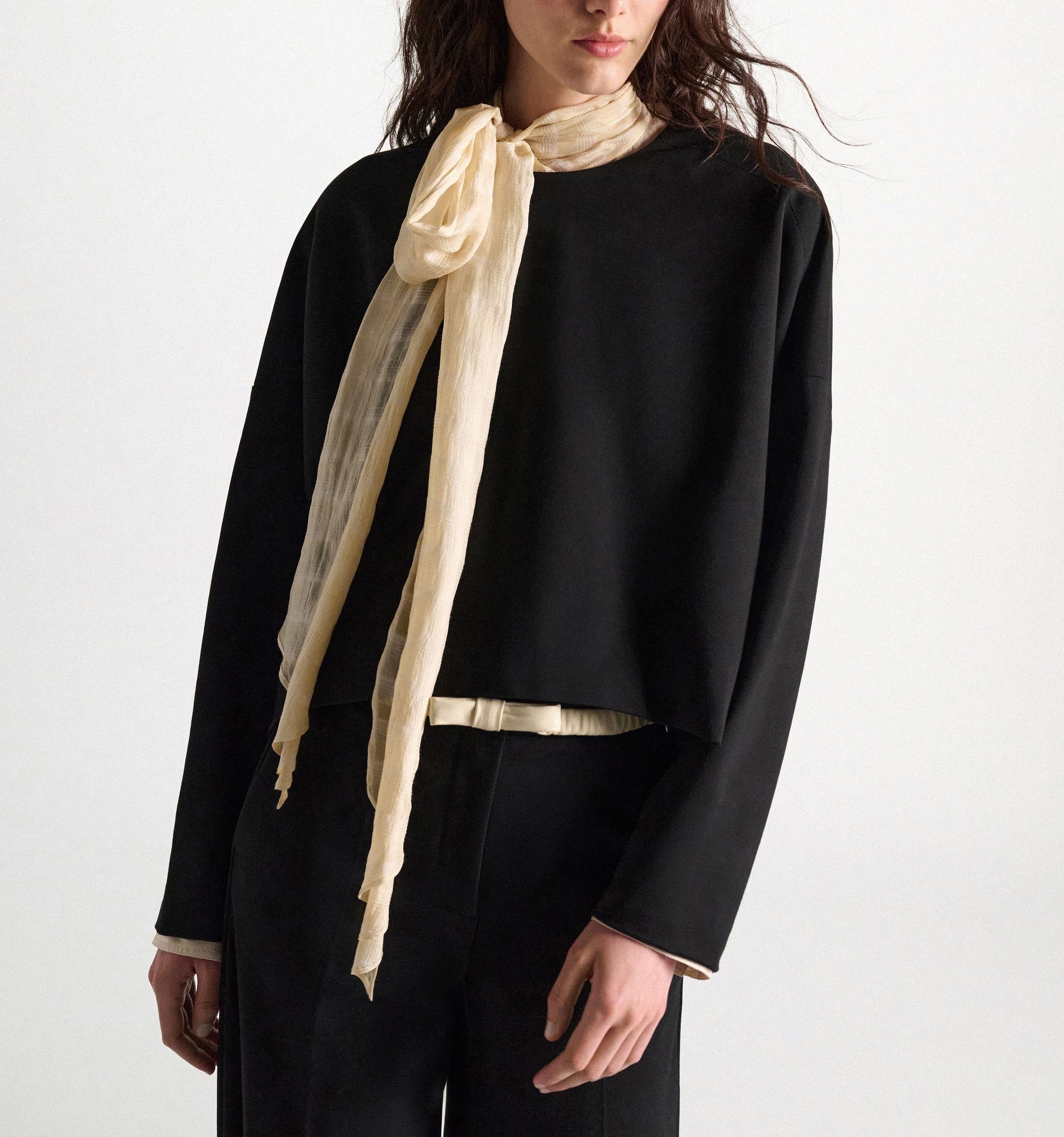 A person stands against a plain background wearing the Dorothee Schumacher Emotional Essence Blouse—an oversized black long-sleeve blouse paired with pants. A light beige sheer scarf is tied in a bow closure around their neck.