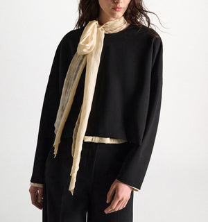 A person stands against a plain background wearing the Dorothee Schumacher Emotional Essence Blouse—an oversized black long-sleeve blouse paired with pants. A light beige sheer scarf is tied in a bow closure around their neck.