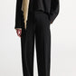 A person wearing Dorothee Schumacher Emotional Essence Pants in loose black Punto Milano fabric with an unfinished hemline, paired with a matching black long-sleeve top, black pointed shoes, and a light beige scarf, stands against a plain white background.