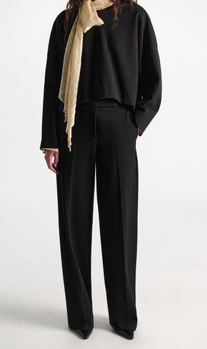 A person wearing Dorothee Schumacher Emotional Essence Pants in loose black Punto Milano fabric with an unfinished hemline, paired with a matching black long-sleeve top, black pointed shoes, and a light beige scarf, stands against a plain white background.