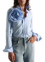 A person wearing the Dorothee Schumacher Striped Power Blouse—a light blue cotton poplin shirt featuring stripes, a large fabric flower on one shoulder, and flared cuffs—paired with light wash jeans.