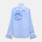 Dorothee Schumacher Striped Power Blouse in light blue cotton poplin, featuring pinstripes, a large fabric rose brooch on the left chest, and ruffled cuffs, displayed on a plain background.