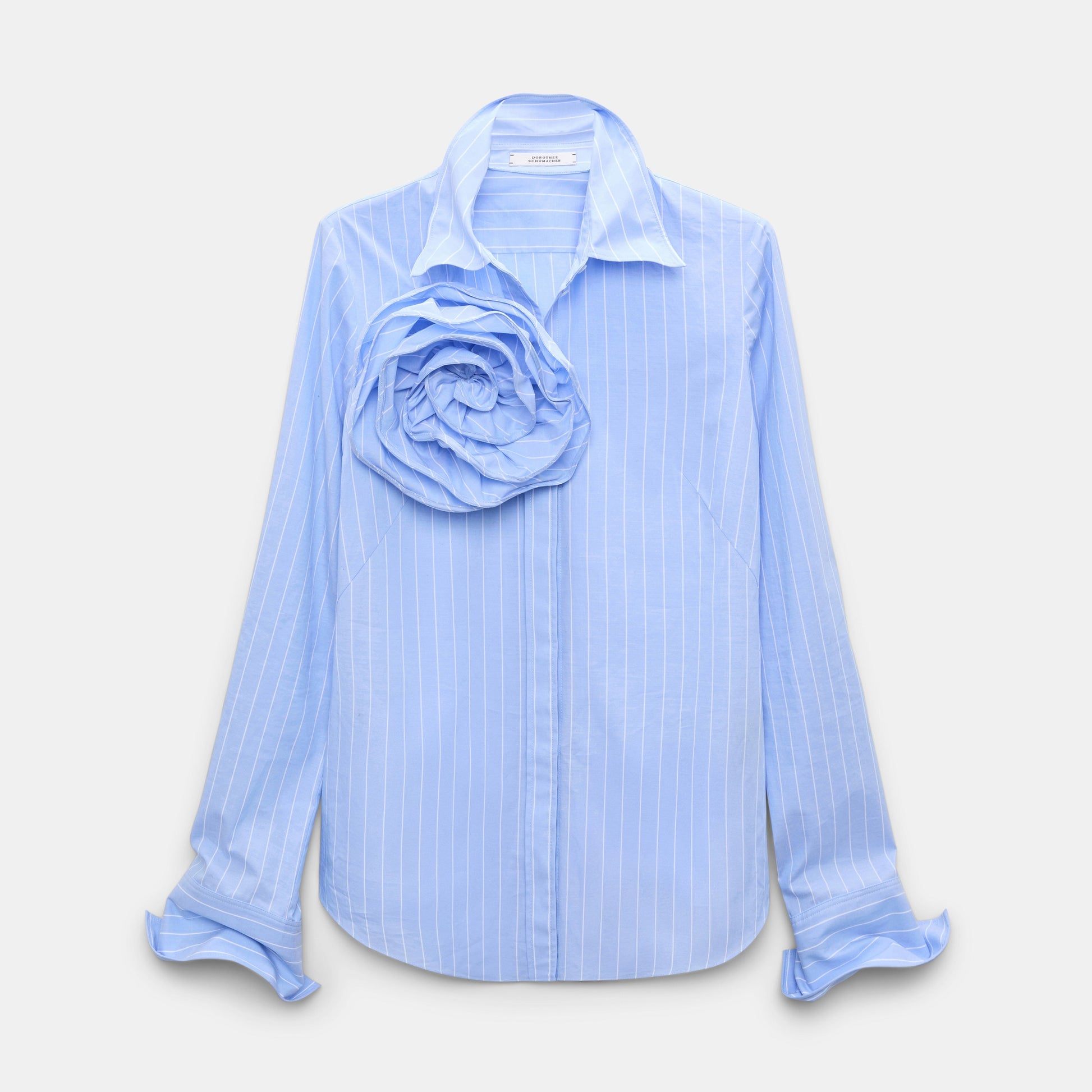 Dorothee Schumacher Striped Power Blouse in light blue cotton poplin, featuring pinstripes, a large fabric rose brooch on the left chest, and ruffled cuffs, displayed on a plain background.