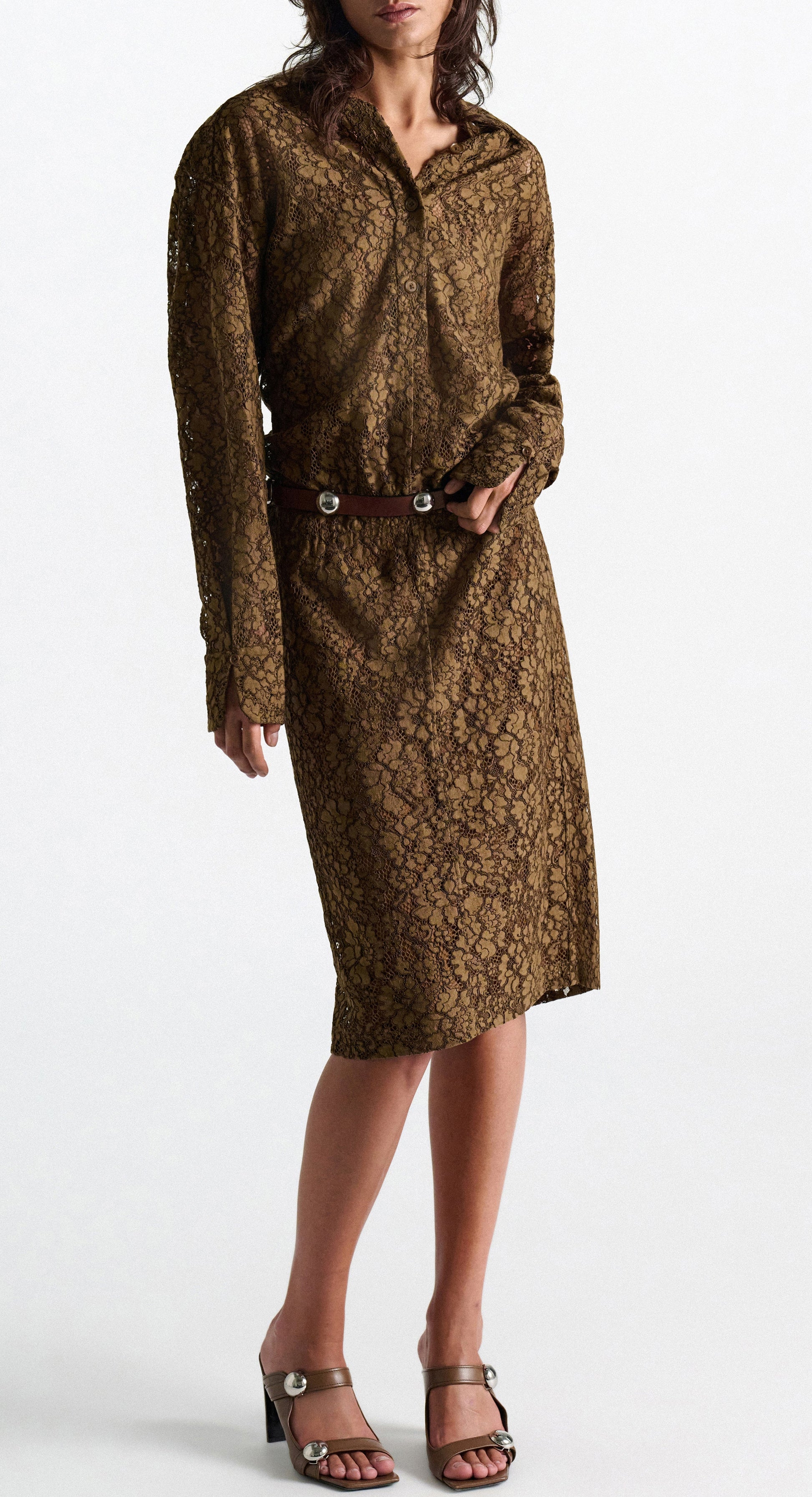 A woman wearing the DS Powerful Lace Dress by Dorothee Schumacher—a brown guipure lace shirtdress featuring long sleeves, a collar, and a dark belt—pairs it with taupe heeled sandals as she stands against a plain white background.