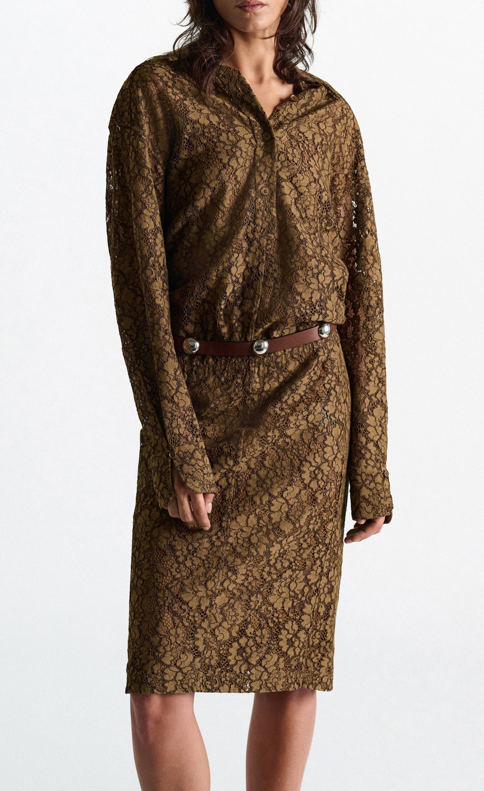 A person wearing the DS Powerful Lace Dress by Dorothee Schumacher—a long-sleeved, brown guipure lace shirtdress with a collar and a brown belt featuring metallic accents—stands against a plain background.
