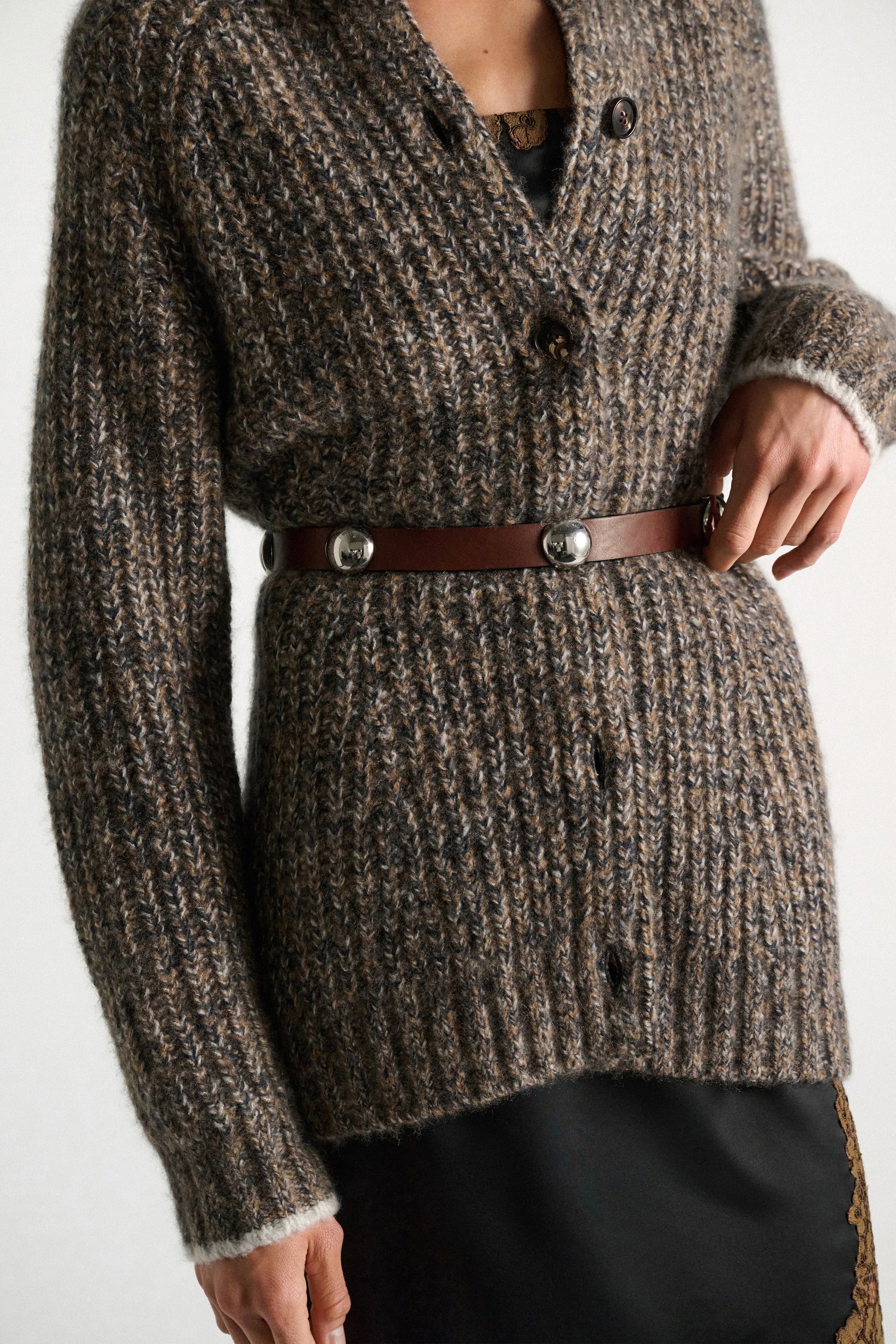 Person wearing a chunky, brown knit cardigan with buttons, belted at the waist with the Dorothee Schumacher True Cuoio Stud Belt—a thin belt featuring a stylish pin stud closure—and a black skirt underneath.