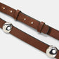 Close-up of two brown leather belts by Dorothee Schumacher: one is the True Cuoio Stud Belt, crafted from soft calfskin with silver metal jingle bells attached; the other is a thin belt featuring a simple buckle and adjustment holes.