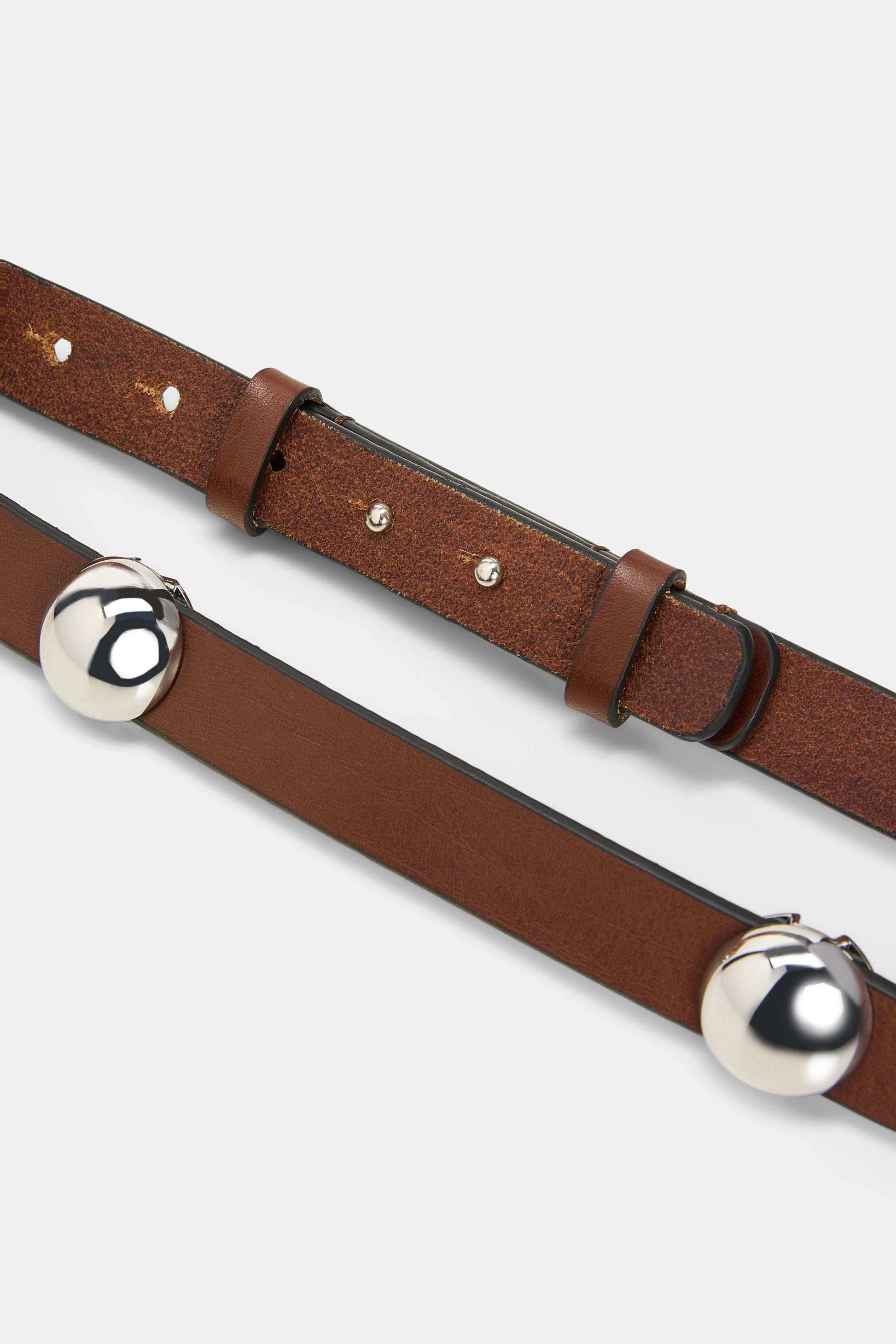 Close-up of two brown leather belts by Dorothee Schumacher: one is the True Cuoio Stud Belt, crafted from soft calfskin with silver metal jingle bells attached; the other is a thin belt featuring a simple buckle and adjustment holes.