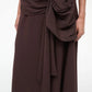 A person wearing the Staud Davina Dress—a dark brown, asymmetrical midi skirt with a large draped bow at the waist and a subtle handkerchief hem—stands against a plain light background.