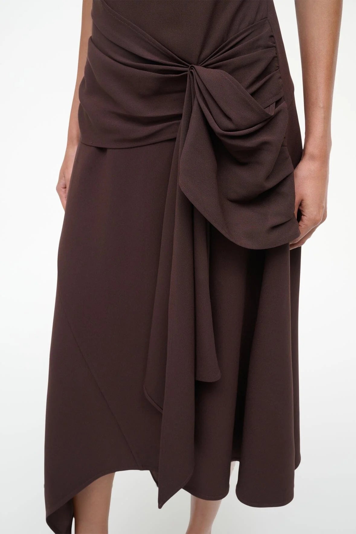 A person wearing the Staud Davina Dress—a dark brown, asymmetrical midi skirt with a large draped bow at the waist and a subtle handkerchief hem—stands against a plain light background.