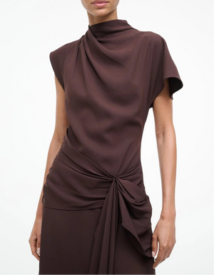 A person wearing the Staud Davina Dress—a short-sleeved, high-neck brown style with draped fabric, a knotted waist detail, and an elegant handkerchief hem—perfect for day-to-night wear.