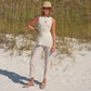 A woman stands on white sand in light-colored pants, a sleeveless top, sun hat, sunglasses, and the Carissa Suede Mule Penny by Magnanni, with tall grass and greenery in the background.