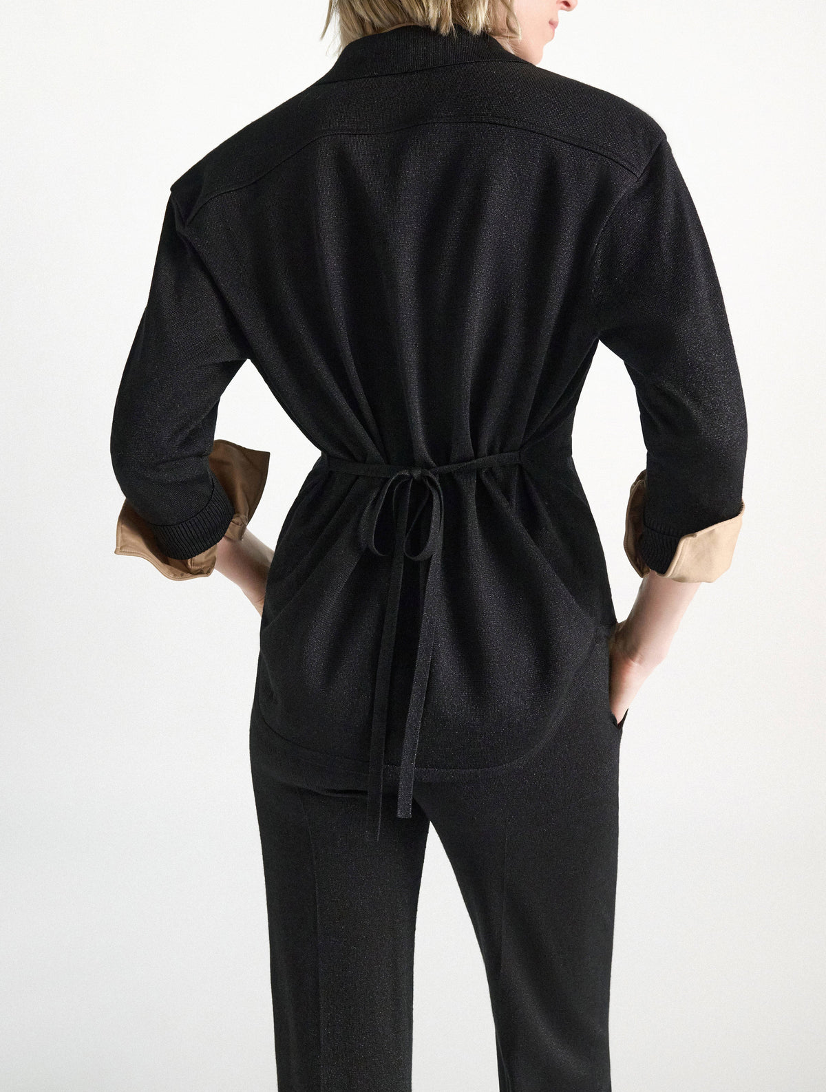 Back view of a person wearing a shimmering Merino-Lurex Shimmering Merino Cardigan with a self-tie sash and rolled tan cuffs, paired with matching black trousers.
