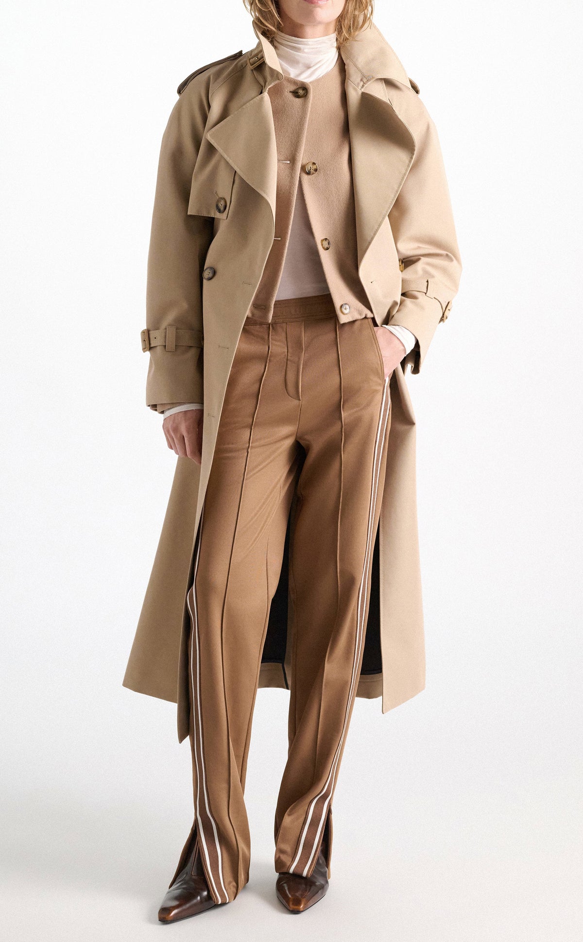 A person wearing a tan trench coat, a neutral-toned quilted knit cardigan, and Dorothee Schumacher Everyday Cool Pants in brown with white side stripes and side slits at the hem.