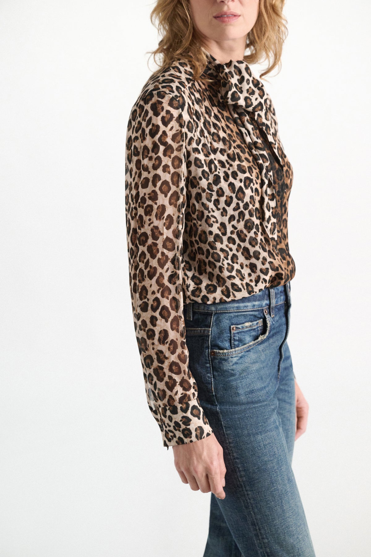 A woman wearing the Dorothee Schumacher Wild at Heart Blouse in leopard print viscose georgette with a high-collar tie-neck detail tucked into blue jeans.