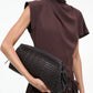 A woman in a brown short-sleeve dress holds the Staud Maude Woven Soft Clutch under her arm against a plain background.