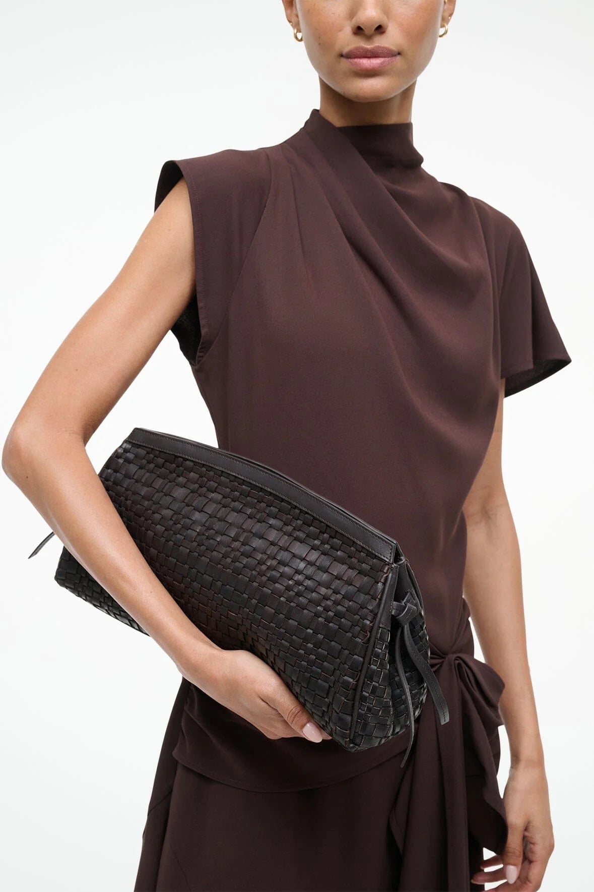 A woman in a brown short-sleeve dress holds the Staud Maude Woven Soft Clutch under her arm against a plain background.