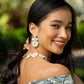 A woman smiles outdoors, wearing Juliska Capucine De Wulf's Mermaid Garden Convertible Floral Post Drop Earrings with a matching necklace and an off-the-shoulder top, set against lush greenery.