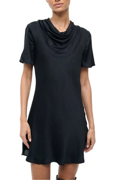 A woman wearing the Staud Iris Silk Dress, a black mini dress with a cowl neck, stands against a white background.