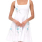 A woman wears the Staud Mini Wells Dandelion Dress—a sleeveless white cotton poplin style with a square neckline, blue floral embroidery, and corset-style paneling—standing with her hands clasped.
