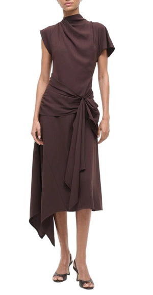 A person wearing the Staud Davina Dress, a sleeveless dark brown asymmetrical day-to-night dress with draped fabric and a waist tie, paired with black open-toe heels.