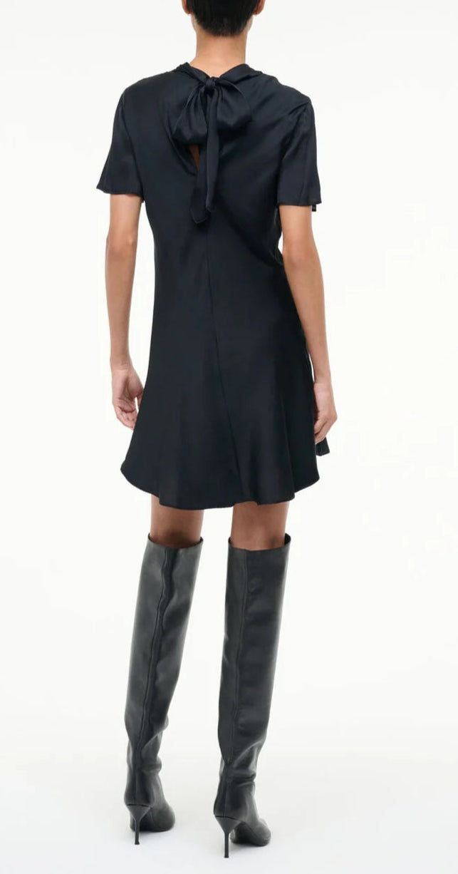A person stands facing away, wearing the Staud Iris Silk Dress—a short-sleeved black mini dress with a back tie detail—paired with knee-high black heeled boots.