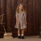 A young girl with curly hair smiles while standing on a rug next to a stack of books, a wooden chair, and a wood-paneled wall. She is wearing the Rylee & Cru Girls’ India Dress, which features a patterned design, smocked bodice, and mini skirt. She completes her outfit with a headband and black shoes.