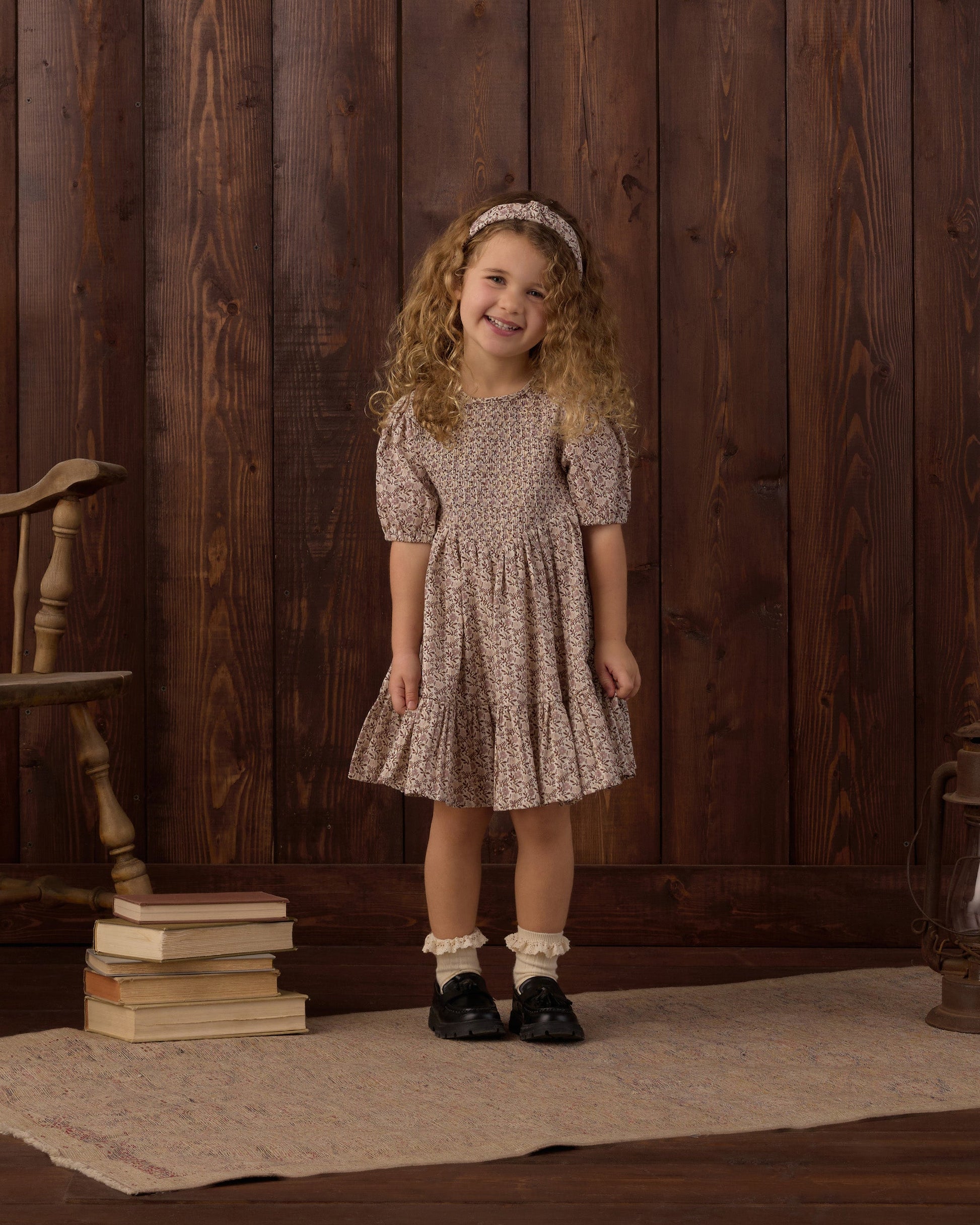 A young girl with curly hair smiles while standing on a rug next to a stack of books, a wooden chair, and a wood-paneled wall. She is wearing the Rylee & Cru Girls’ India Dress, which features a patterned design, smocked bodice, and mini skirt. She completes her outfit with a headband and black shoes.