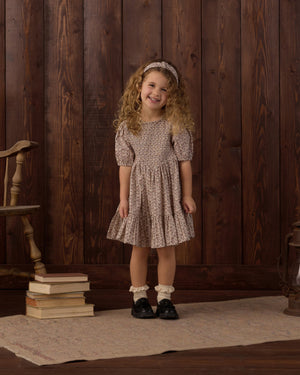 A young girl with curly hair smiles while standing on a rug next to a stack of books, a wooden chair, and a wood-paneled wall. She is wearing the Rylee & Cru Girls’ India Dress, which features a patterned design, smocked bodice, and mini skirt. She completes her outfit with a headband and black shoes.
