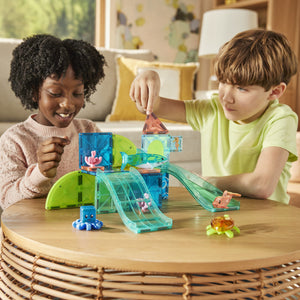 Two children sit at a round table, playing with the Magnatiles Coral Reef 25-Piece Set, building an imaginative coral reef adventure with colorful magnetic tiles, ramps, slides, and small ocean animal figures.