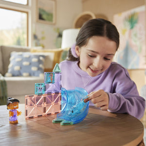 A girl in a purple sweatshirt plays with her Magnatiles Dolphin Bay 13-Piece Set and a small slide on a wooden table in the living room.