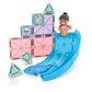 A child figurine stands at the top of a blue slide, with a dolphin toy figure at the bottom, all nestled among MAGNA-TILES® Dolphin Bay 13-Piece Set translucent magnetic tiles featuring sea creature designs by Magnatiles.