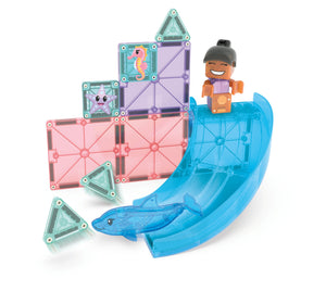 A child figurine stands at the top of a blue slide, with a dolphin toy figure at the bottom, all nestled among MAGNA-TILES® Dolphin Bay 13-Piece Set translucent magnetic tiles featuring sea creature designs by Magnatiles.