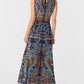 A woman is shown from the back wearing the Vivienne Brise Dress by Emporio Sirenuse, a sleeveless, floor-length piece with blue, brown, and orange patterns, Brisé Cutwork detailing, and a sheer upper back panel.