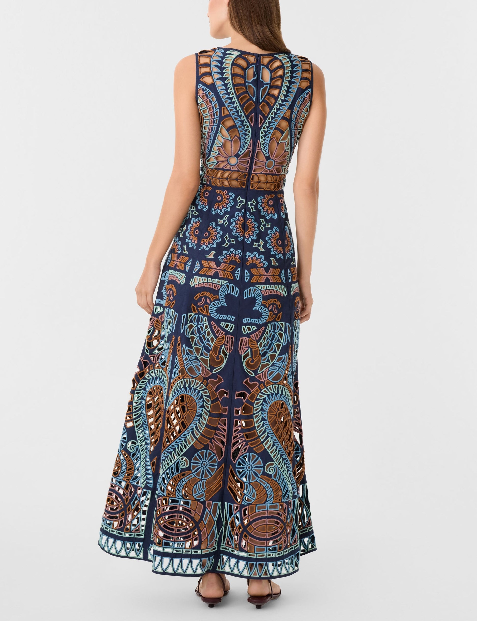 A woman is shown from the back wearing the Vivienne Brise Dress by Emporio Sirenuse, a sleeveless, floor-length piece with blue, brown, and orange patterns, Brisé Cutwork detailing, and a sheer upper back panel.