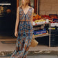 A woman in a patterned Emporio Sirenuse Vivienne Brise Dress stands by a street market with fruit boxes behind her, holding a woven bag.