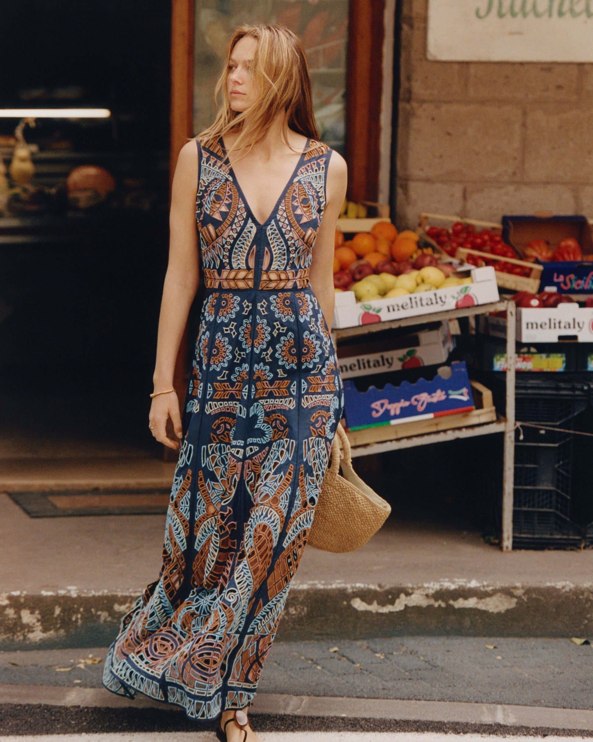 A woman in a patterned Emporio Sirenuse Vivienne Brise Dress stands by a street market with fruit boxes behind her, holding a woven bag.