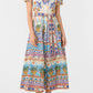 A woman wears the Emporio Sirenuse Giselle Chimera Dress, a short-sleeve, button-up shirt dress with a vibrant Indonesian-inspired print, cinched with a matching belt and paired with tan sandals.