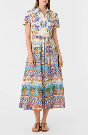 A woman wears the Emporio Sirenuse Giselle Chimera Dress, a short-sleeve, button-up shirt dress with a vibrant Indonesian-inspired print, cinched with a matching belt and paired with tan sandals.