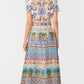 A woman stands facing away, wearing the Emporio Sirenuse Giselle Chimera Dress—an ankle-length, short-sleeve piece with a vibrant Chimera Print inspired by Indonesian textiles, paired with sandals.