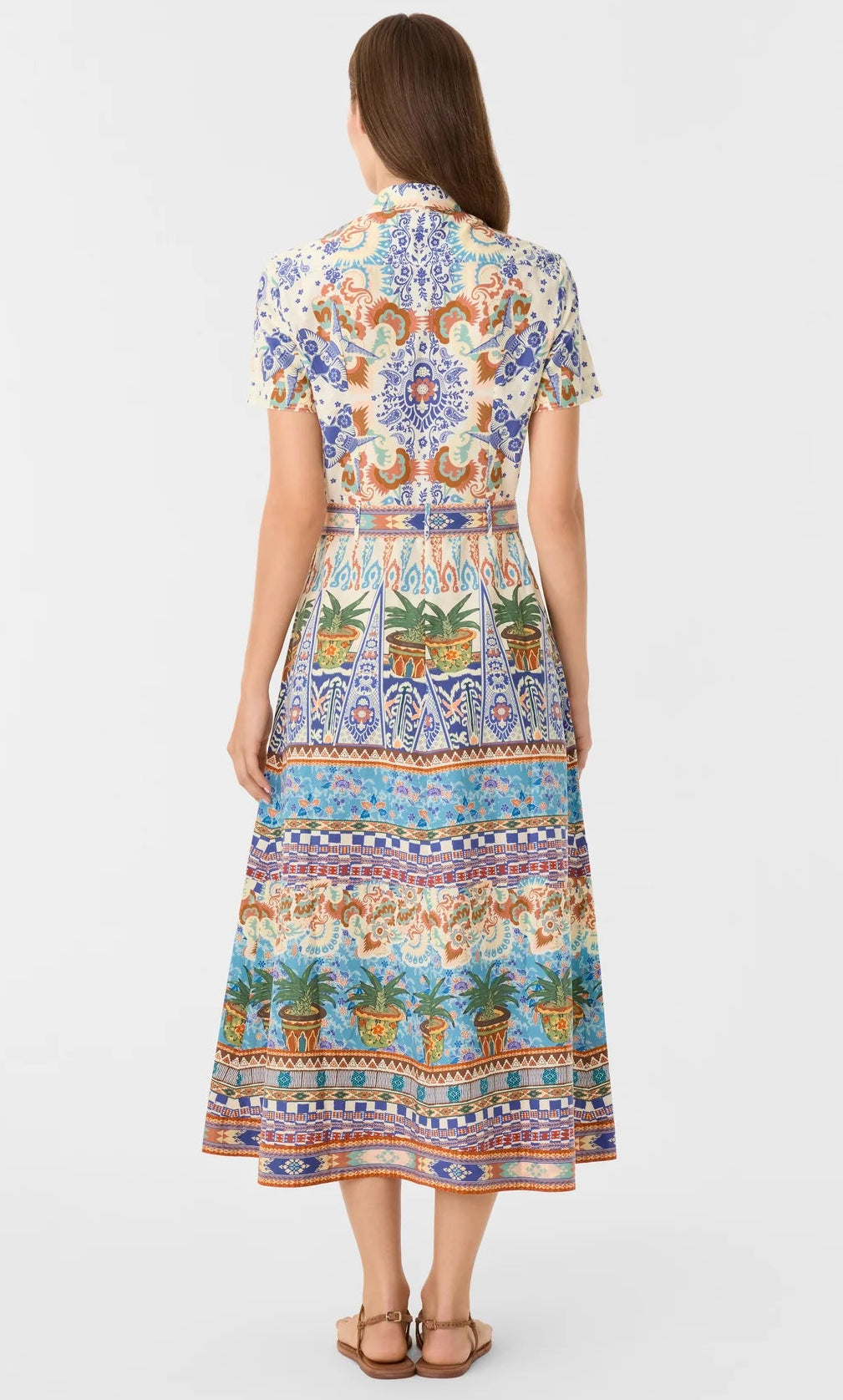 A woman stands facing away, wearing the Emporio Sirenuse Giselle Chimera Dress—an ankle-length, short-sleeve piece with a vibrant Chimera Print inspired by Indonesian textiles, paired with sandals.