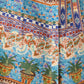 The Giselle Chimera Dress by Emporio Sirenuse features colorful horizontal bands with floral motifs, geometric designs, and potted plant illustrations in blue, orange, green, and brown tones inspired by Indonesian textiles.