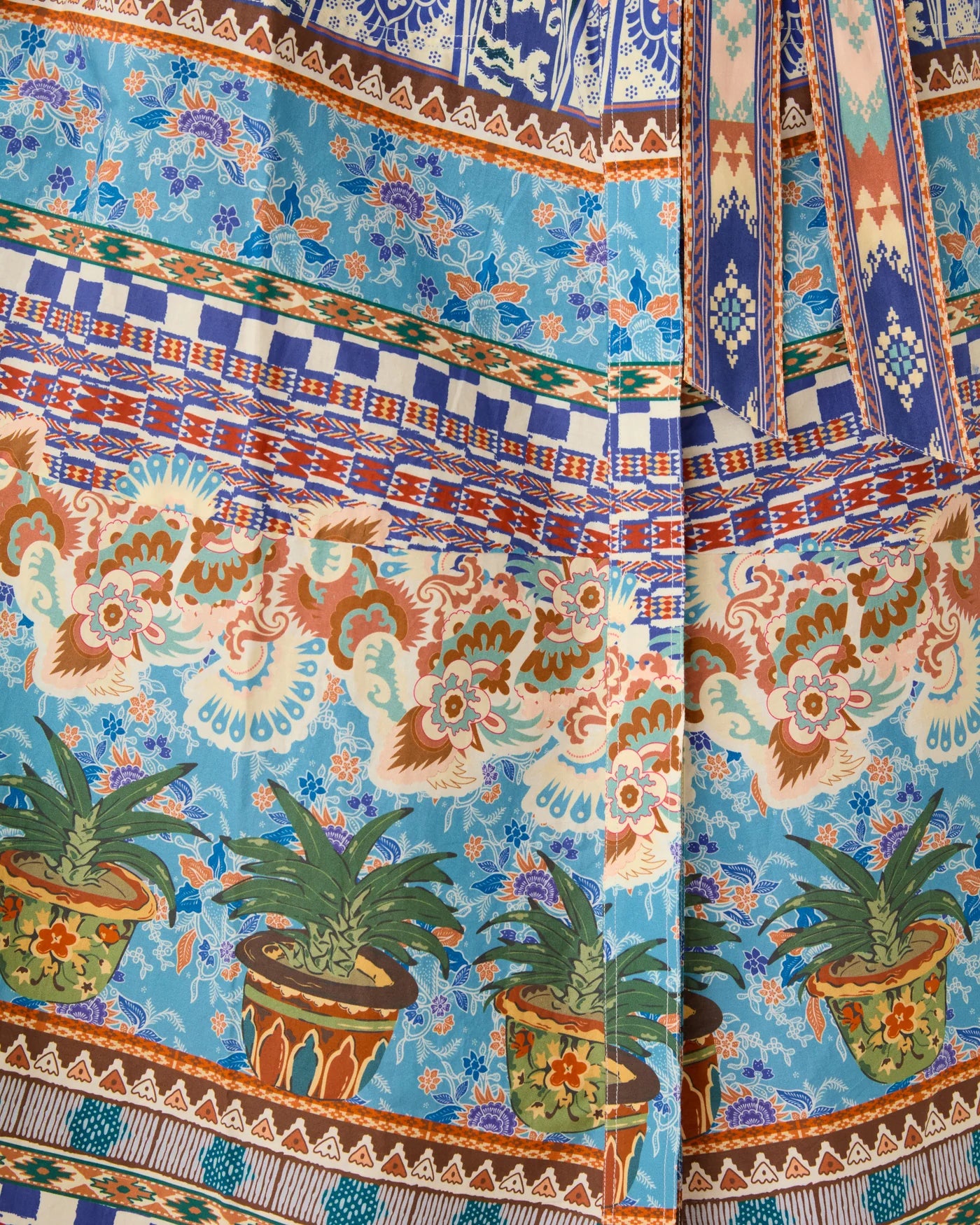 The Giselle Chimera Dress by Emporio Sirenuse features colorful horizontal bands with floral motifs, geometric designs, and potted plant illustrations in blue, orange, green, and brown tones inspired by Indonesian textiles.