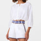 Woman wearing an Emporio Sirenuse New Jinny Blue Horizon Top, a white linen cropped style with blue embroidered patterns, paired with high-waisted white shorts featuring matching blue embroidery.